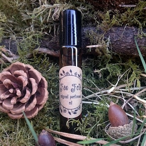 Puede incluir: Un frasco de vidrio marrón con tapa negra y una etiqueta de papel marrón que dice "Fae Folk original perfumed oil". El frasco está sobre un lecho de musgo verde.