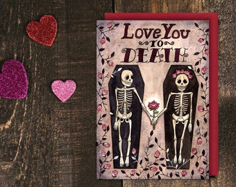 Può includere: Un biglietto di San Valentino con uno sfondo scuro che presenta due scheletri che si tengono per mano in bare. Il biglietto recita "Love You To Death" con una rosa tra gli scheletri.
