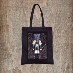 May include: A black tote bag featuring a rectangular graphic print of a stylized nutcracker figure. The nutcracker wears a blue hat and uniform with white hair and beard, holding a staff topped with a skull, set against a dark, branch-like background.