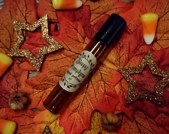 May include: A brown glass bottle with a black rollerball top. The bottle has a white label with the text "Sleepy Pumpkins" and "original perfumed oil". The bottle is surrounded by autumnal decorations, including orange and yellow leaves, candy corn, and gold glitter stars.