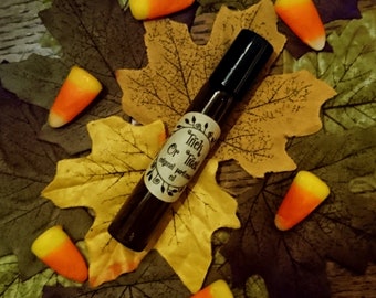 May include: A brown glass bottle with a black cap and a white label that says "Trick or Treat" original perfume oil. The bottle is surrounded by yellow and green autumn leaves and candy corn.