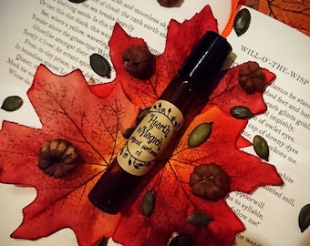 Può includere: Un piccolo flacone di vetro marrone con un'etichetta che recita "Hearth Magick" e "olio profumato originale". Il flacone è appoggiato su una foglia di acero rossa con piccoli semi marroni e verdi sparsi intorno. Il flacone è sopra un libro con il testo "Will-O'-The-Wisp" visibile.