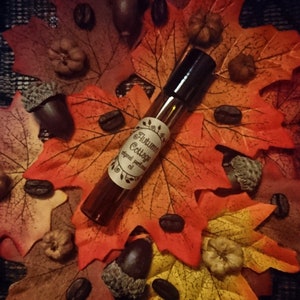 Peut inclure: Un petit flacon en verre brun avec un bouchon noir, contenant une huile de parfum. Le flacon est étiqueté "Autumn Cottage Original Perfume Oil". Le flacon est entouré de feuilles d'automne, de glands et de grains de café.