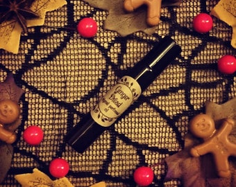 Puede incluir: Una pequeña botella de vidrio de aceite esencial "Ginger Bread" con una etiqueta negra y un diseño de telaraña blanca. La botella está rodeada de caramelos rojos, hombres de jengibre y hojas de otoño.