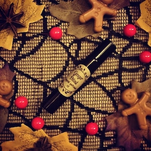 Puede incluir: Una pequeña botella de vidrio de aceite esencial "Ginger Bread" con una etiqueta negra y un diseño de telaraña blanca. La botella está rodeada de caramelos rojos, hombres de jengibre y hojas de otoño.