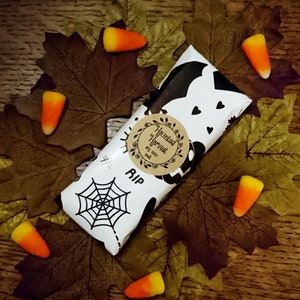 May include: A white and black Halloween-themed soy wax melt bar with a spiderweb and the words "RIP" and "Haunted Harvest". The bar is surrounded by fall leaves and candy corn.