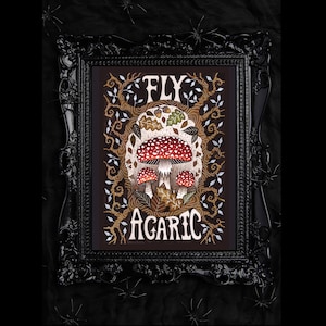 K&ouml;nnte beinhalten: Eine gerahmte Illustration eines roten und wei&szlig;en Pilzes mit dem Text "FLY AGARIC" in einer stilisierten Schriftart. Der Pilz ist von Bl&auml;ttern und &Auml;sten umgeben.