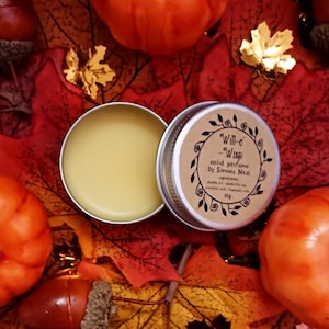 Op de afbeelding: Een klein blikje met vaste parfum genaamd "Will-o'-Wisp" van Simons Nest. Het blikje is omringd door herfstbladeren en pompoenen. Het etiket luidt "Will-o'-Wisp solid perfume by Simons Nest. Ingredients: jojoba oil, candelilla wax, essential oils, fragrance oil. 10g."