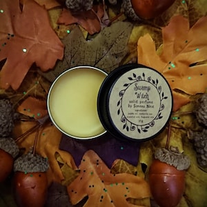 Puede incluir: Un pequeño bote redondo abierto de perfume sólido "Swamp Witch" de Simons Nest. El bote está sobre un lecho de hojas de otoño y bellotas. El perfume es de color amarillo claro.