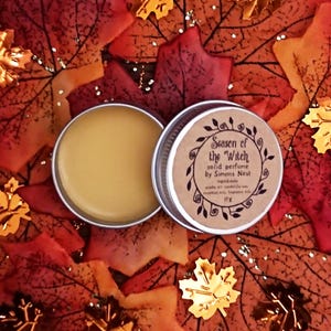 May include: A silver tin of solid perfume, open to reveal a yellow balm. The lid, next to the tin, has the text "Season of the Witch" and "solid perfume by Simons Nest". The background is autumnal leaves.