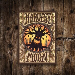 May include: An art print with the words "HARVEST MOON" in a decorative font. The image shows a tree with a carved pumpkin face, surrounded by wheat and a brown border. The colours are orange, brown, and beige.
