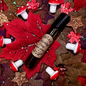 Peut inclure: Un flacon en verre brun avec une étiquette en papier brun qui indique "Nutcracker Creep Original Perfumed Oil". Le flacon est entouré de feuilles d'automne rouges et vertes et de champignons blancs.