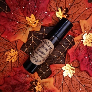 Peut inclure: Un flacon roll-on noir étiqueté "Gloomy Sweater original perfumed oil" repose sur un lit de feuilles d'automne. Les feuilles sont rouges, oranges et dorées, avec des accents de feuilles d'or décoratives.