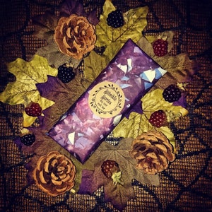 May include: A purple and blue soy wax melt bar with a brown label that says "Bramble Berries Soy Wax Melt Bar". The bar is surrounded by fall leaves, pine cones, and blackberries.