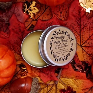 May include: A small tin of solid perfume with a brown label that reads "Pumpkin Patch Pieme solid perfume by Simons Nest". The tin is open and the perfume is a pale yellow color. The tin is surrounded by fall leaves.