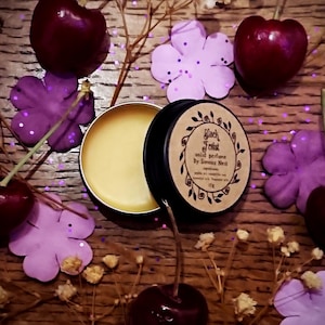 May include: A small, round tin of solid perfume called "Black Forest" by Simmias Neil. The tin is open, revealing a yellow, waxy perfume inside. The tin is surrounded by purple flowers and red cherries.