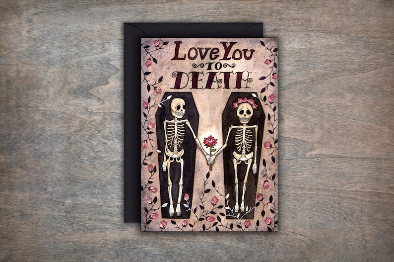 Puede incluir: Una tarjeta de felicitaci&oacute;n con una ilustraci&oacute;n de acuarela de dos esqueletos tomados de la mano en ata&uacute;des. Los esqueletos llevan coronas de flores y el texto "Love You To Death" est&aacute; escrito encima de ellos.