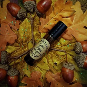 May include: A roll-on bottle of "Green Man" original perfumed oil is positioned on a bed of autumn leaves and acorns. The bottle has a black cap and a label with a floral design. The background features fall foliage in shades of yellow, orange, and brown.