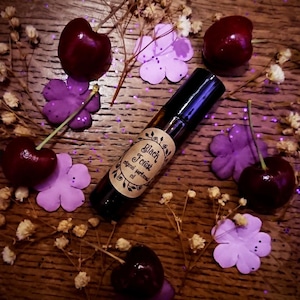 Könnte beinhalten: Eine kleine braune Glasflasche mit einem schwarzen Roll-on-Applikator. Die Flasche hat ein Etikett mit der Aufschrift "Black Forest original perfume oil". Die Flasche ist von lila und weißen Blumen sowie roten Kirschen umgeben.
