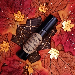 以下が含まれることがあります： 「Pumpkin Spice Latte」と書かれたラベルが付いた黒いロールオンボトルが、秋の葉のベッドの上に置かれています。葉は赤、オレンジ、金の色合いで、金の葉のアクセントが付いています。ボトルには透明な液体が入っています。