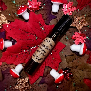 Op de afbeelding: Een klein bruin glazen flesje met een etiket waarop staat "Sugarplum Visions Original Perfume Oil". Het flesje is omringd door rode en groene herfstbladeren en witte paddenstoelen.