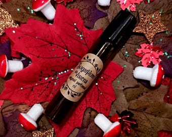 May include: A small brown glass bottle with a label that reads "Sugarplum Visions Original Perfume Oil". The bottle is surrounded by red and green fall leaves and white mushrooms.