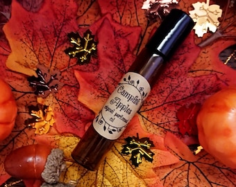 Puede incluir: Una botella de vidrio marrón con una tapa negra y una etiqueta que dice "Campfire Apples Original Perfume Oil". La botella está rodeada de hojas de otoño y bellotas.