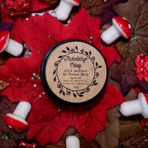 Könnte beinhalten: Eine kleine, runde Dose mit festem Parfüm mit der Aufschrift "Nutcracker Creep" von Simons Nest. Die Dose ist mit einer fantasievollen Illustration eines Nussknackers und den Worten "festes Parfüm" und "Zutaten" verziert. Die Dose ist von roten und braunen Herbstblättern und weißen Keramikpilzen umgeben.