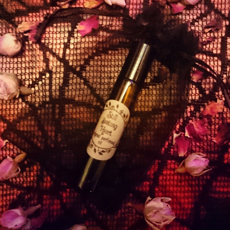 May include: A small brown glass bottle with a black cap and a white label that reads "Still Beating Heart  original perfume oil". The bottle is sitting on a black lace fabric with pink rose petals.