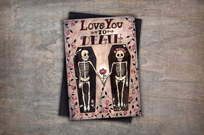 Puede incluir: Una tarjeta de felicitaci&oacute;n con una ilustraci&oacute;n de acuarela de dos esqueletos tomados de la mano en ata&uacute;des rodeados de rosas rosas. El texto de la tarjeta dice "Love You To Death."