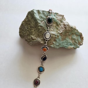 May include: Silver bracelet featuring seven oval-shaped gemstones in various colors: green, white, orange, dark blue, turquoise, and maroon. The bracelet is displayed on a textured, green-gray rock against a white background.