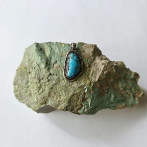 May include: A turquoise pendant with a silver-colored frame and bail. The pendant has an oval shape and is set against a green and gray stone. The turquoise stone is a bright blue color.