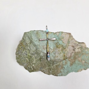May include: A silver and gold cross pendant with turquoise and red accents. The cross is displayed on a textured, green-gray stone. The pendant has a loop at the top for a chain.