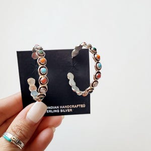 May include: A pair of silver hoop earrings with colorful stones. The earrings are on a black card with the text "INDIAN HANDCRAFTED STERLING SILVER."