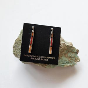 May include: A pair of sterling silver earrings with a rectangular design. The earrings feature a red, orange, and yellow inlay with a beaded border. The earrings are displayed on a black card with the text "GENUINE INDIAN HANDCRAFTED STERLING SILVER".