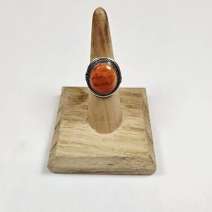 May include: A silver ring with an oval, orange-colored stone. The stone is set in a silver bezel with a decorative border. The ring is displayed on a wooden ring holder against a white background. The ring is a piece of jewelry.