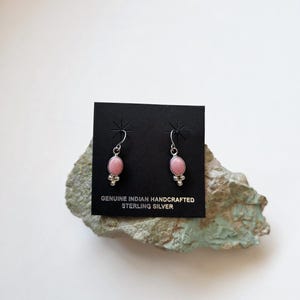 May include: A pair of sterling silver dangle earrings featuring oval pink stones and small silver bead accents. The earrings are attached to a black display card with the text GENUINE INDIAN HANDCRAFTED STERLING SILVER printed in gold.