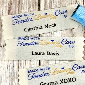 May include: Three white fabric labels with blue and gold embroidery. The labels read "Made with Tender Loving Care By" followed by a name. The names are Cynthia Neck, Laura Davis, and Grama XOXO.