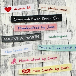 May include: A collection of colorful fabric labels with various text, including "Auntie M", "Savannah River Boot Co.", "Handcrafted by Jane", "MAIDS A MAKIN", "Nan loves me", "madebyjane", "Save a Tree USE ME!", "Handcrafted by Gwyn", and "Sew Simple by Beth".