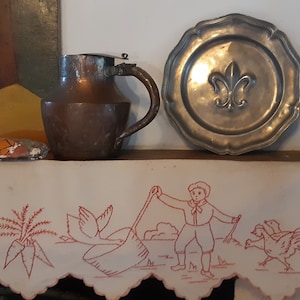 Könnte beinhalten: Ein antiker Kupferkrug mit Deckel und Henkel steht auf einem Regal mit einem Zinnteller, der ein Fleur-de-lis-Design zeigt. Unter dem Regal befindet sich ein weißer Stoff mit roten Stickereien, die einen Jungen mit einer Harke, einen Hahn und einen Bund Karotten darstellen.