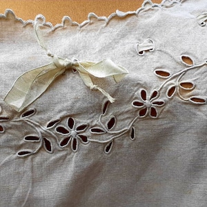 May include: A white linen fabric with a scalloped edge and a delicate floral design. A white satin ribbon is tied in a bow at the top.