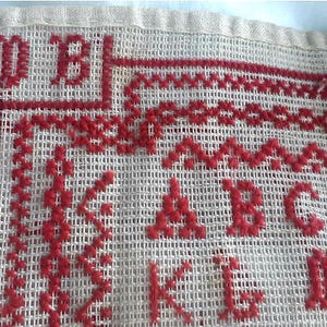 以下が含まれることがあります： ヴィンテージのクロスステッチサンプラーの一部をクローズアップ。赤色の刺繍文字、数字、装飾ステッチがクリーム色の生地に施されています。テキストには、ABC、K、L、Dの文字が含まれています。