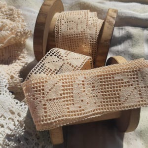 Op de afbeelding: Close-up van beige kanten lintspoelen. Het lint is gemaakt van een delicate, open weefsel met een herhalend geometrisch patroon. De spoelen zijn van hout en zijn gedeeltelijk zichtbaar. Andere kant is zichtbaar op de achtergrond.