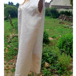 May include: A white linen sleeveless dress with a lace trim around the neckline. The dress is on a mannequin in a garden setting.