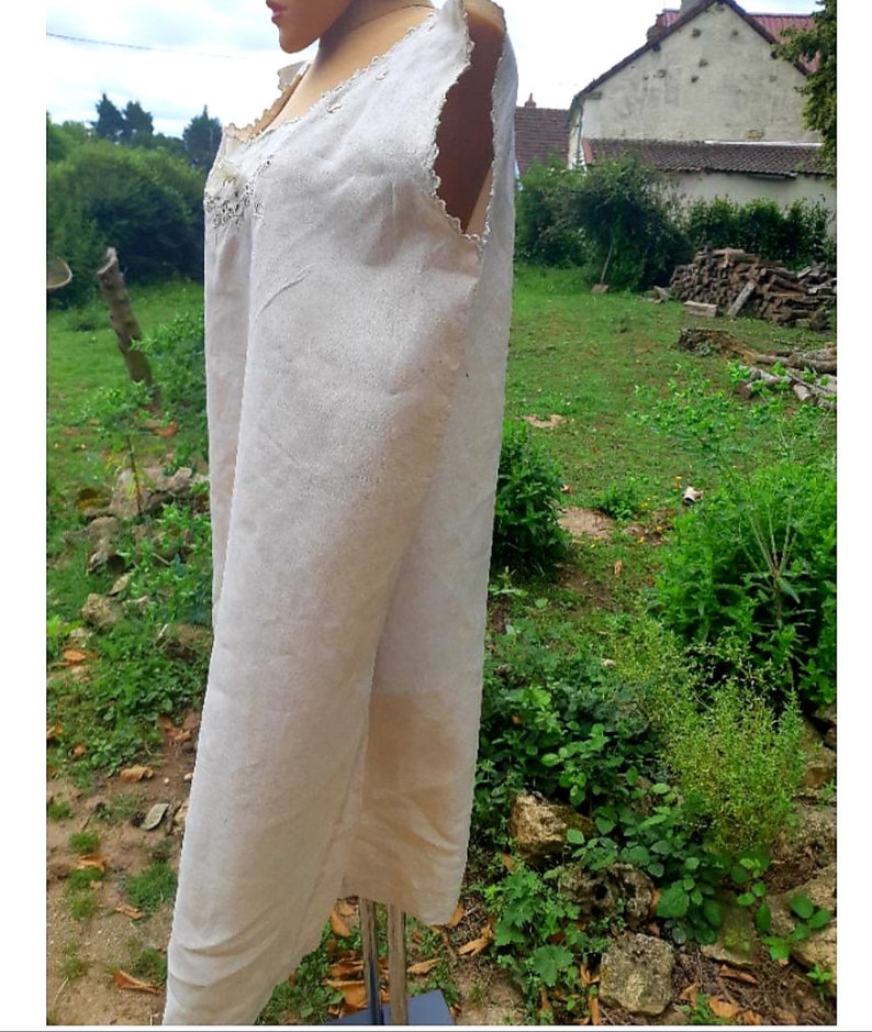 May include: A white linen sleeveless dress with a lace trim around the neckline. The dress is on a mannequin in a garden setting.