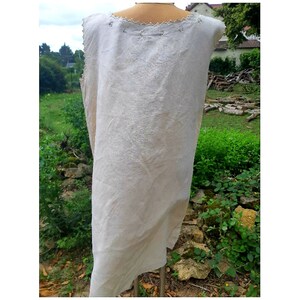 May include: A sleeveless, off-white linen tunic with a scalloped neckline and decorative stitching. The tunic is displayed on a mannequin against a backdrop of green foliage.