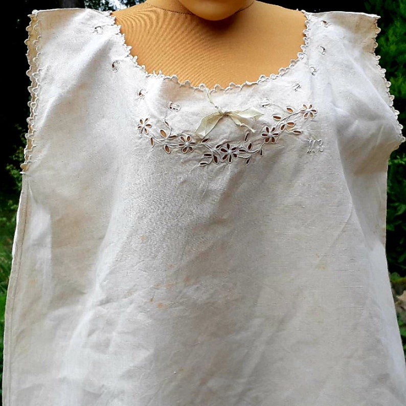 May include: A white sleeveless cotton top with delicate lace trim and embroidered floral details. The top has a scalloped neckline and a bow at the center.