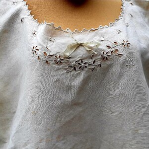 May include: White cotton blouse with a delicate lace trim and embroidered floral design. The neckline features a small bow. The blouse has the initials "MR" embroidered on the front.