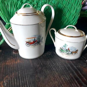 May include: A white porcelain tea set with gold trim, featuring a teapot and a sugar bowl. Each piece is decorated with a colorful illustration of a horse-drawn carriage. The set is placed on a dark wooden surface.
