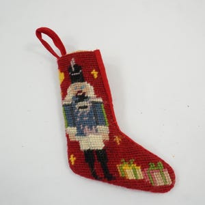 Könnte beinhalten: Ein roter Weihnachtsstrumpf in Stickoptik mit einem zentralen Nussknacker in blauer Uniform, verziert mit kleinen gelben Sternen und zwei Geschenkpaketen an der Spitze. Der Strumpf hat eine rote Stoffschlaufe zum Aufhängen am oberen Rand.